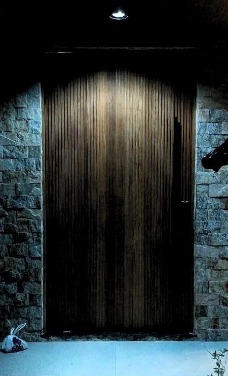 Entrada com porta de madeira iluminada, parede de pedra e escultura ao lado.