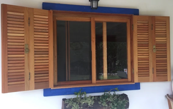 Janela de madeira com venezianas abertas, moldura azul na parede e floreira abaixo.
