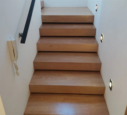 Escada com degraus amadeirados, corrimão preto e luzes na parede lateral.