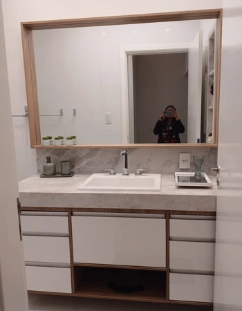 Gabinete branco e amadeirado sob bancada clara e espelho que reflete uma pessoa fotografando.